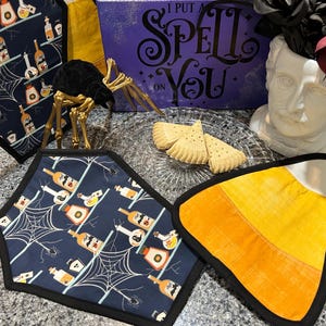 May include: A Halloween-themed still life featuring a variety of items. Includes a navy blue pot holder with a spiderweb and potion bottle design, a candy corn pot holder, and a decorative sign that reads "I PUT A SPELL ON YOU".