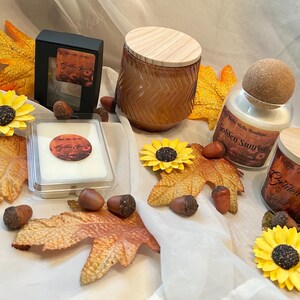 May include: A display of autumn-themed products, featuring a candle jar with a wooden lid, a glass bottle with a wooden stopper, and a small black box. Artificial sunflowers, autumn leaves, and acorns are arranged around the items.