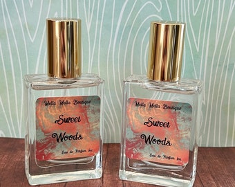 May include: Two clear glass perfume bottles with gold-colored caps. Each bottle has a label that reads "Mella Molla Boutique Sweet Woods Eau de Parfum 1oz". The background is a light blue and white pattern.