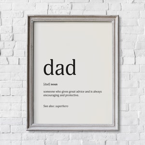May include: Framed print with the word "dad" in a bold, black font. The definition of "dad" is provided below, describing someone who gives great advice and is always encouraging and protective. The phrase "See also: superhero" is at the bottom.