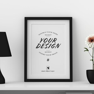 May include: A black framed print with the words "YOUR DESIGN" in a stylish font. A black lamp and a potted flower with a white and peach bloom sit on a white shelf. The image is a mockup.