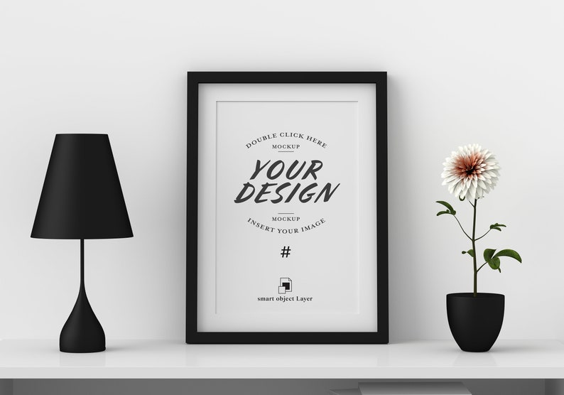 May include: A black framed print with the words "YOUR DESIGN" in a stylish font. A black lamp and a potted flower with a white and peach bloom sit on a white shelf. The image is a mockup.