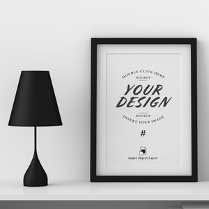 May include: A black framed print with the text "YOUR DESIGN" and a black lamp with a conical shade. The lamp has a slender stem and a rounded base. The print is a mockup with instructions to insert an image.