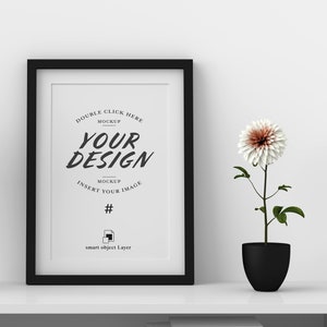May include: A black framed mockup with the text "YOUR DESIGN" and instructions to insert an image. A white and brown flower in a black pot sits to the right of the frame. The frame and pot are on a white shelf.