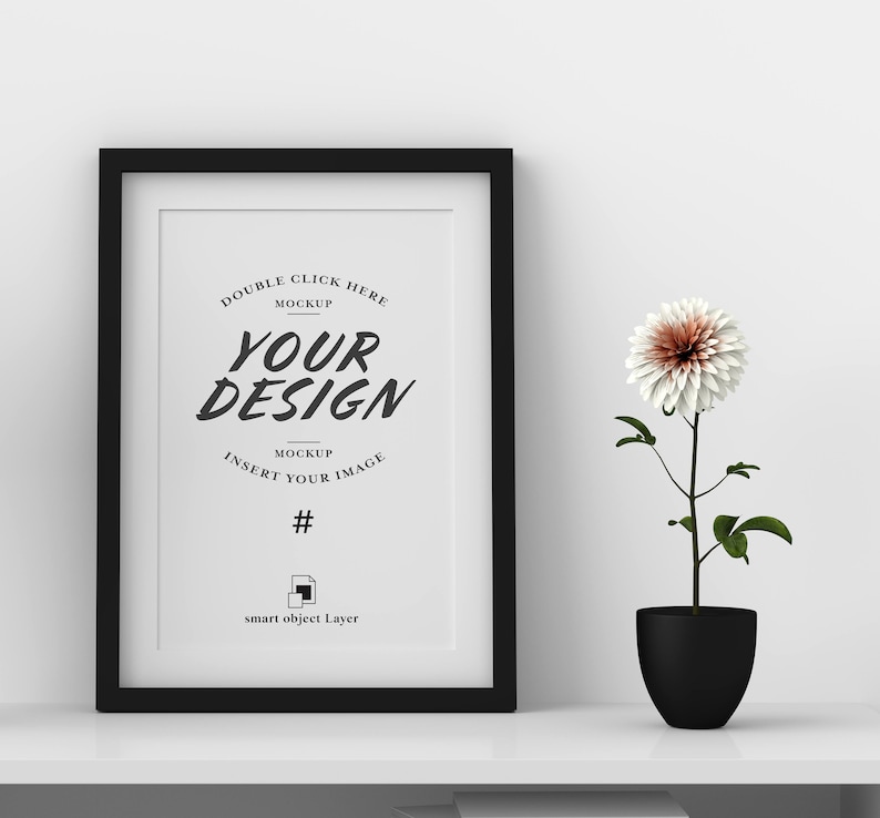 May include: A black framed mockup with the text "YOUR DESIGN" and instructions to insert an image. A white and brown flower in a black pot sits to the right of the frame. The frame and pot are on a white shelf.