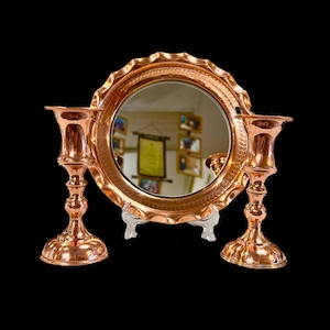 May include: A set of three copper candle holders with a decorative copper mirror in the center. The mirror has a scalloped edge and a hammered finish. The candle holders have a classic design with a round base and a tall, slender stem.