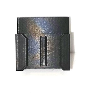 May include: A dark gray, rectangular plastic component featuring a flat top section and a lower base with two vertical, parallel recessed channels centered on the front surface, flanked by two raised, angled side panels on the left and right edges.