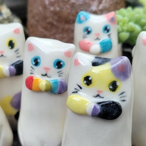 Puede incluir: Seis figuras de cerámica con forma de gato. Cada gato es blanco con detalles coloridos, incluyendo ojos azules, pajaritas arcoíris y manchas de color. Las figuras están de pie sobre una superficie de madera, con un fondo borroso de plantas y cerámica.