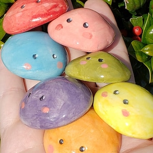 May include: A hand holding a collection of seven small, colourful ceramic mushrooms with painted faces. The mushrooms are arranged in a rainbow order, starting with red and ending with yellow.