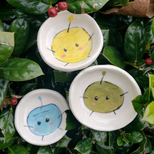 May include: Three small ceramic dishes with hand-painted cartoon characters. The dishes are white with a light blue, yellow, and green character on each. The characters have large eyes and a simple design.