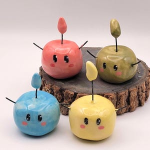 Puede incluir: Cuatro figuras de cerámica coloridas con forma de manzana, con caras pintadas, tallos y pequeños brazos. Las figuras son rojas, verdes, azules y amarillas, y se exhiben sobre una rodaja de madera.