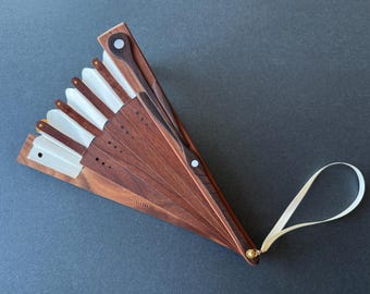 May include: A folding hand fan constructed from dark brown wood slats and off-white fabric panels. The fan features a gold-toned metal pivot point at the base, a light-coloured wrist strap, and small circular white inlays on the wooden frame.