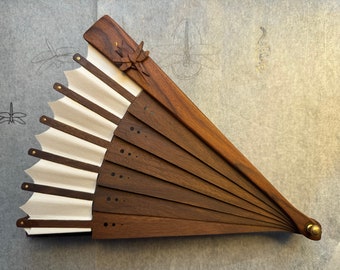 May include: A folding hand fan constructed from dark brown wood slats and off-white fabric. The fan features a carved wooden dragonfly accent on the top slat, small brass rivets connecting the segments, and a brass pivot pin at the base.