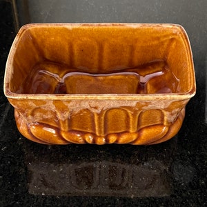 May include: A rectangular, brown ceramic planter with a ribbed exterior and a glossy finish. The planter has a raised interior divider and a curved rim. The color is a warm, earthy tone, suitable for displaying small plants or succulents.