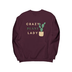 Może przedstawiać: Bordowy sweter z napisem "CRAZY PLANT LADY" w białym i zielonym kolorze. Zielony kaktus jest przedstawiony w brązowej doniczce.
