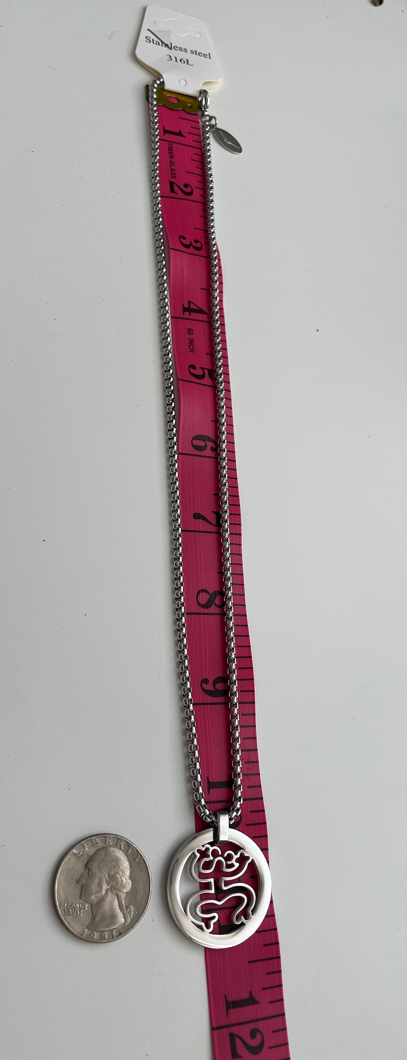 Puede incluir: Un collar de cadena de plata con un colgante redondo que presenta un dise&ntilde;o estilizado. La cadena mide 23 cm de largo y el colgante aproximadamente 2,5 cm de di&aacute;metro. El collar est&aacute; hecho de acero inoxidable.