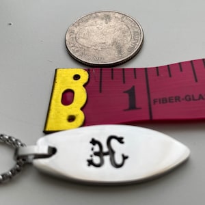 Op de afbeelding: Een zilveren hanger met een zwarte inscriptie van de letters "3C" aan een zilveren ketting. De hanger is ongeveer 2,5 cm lang en ligt naast een liniaal en een Amerikaanse kwart.