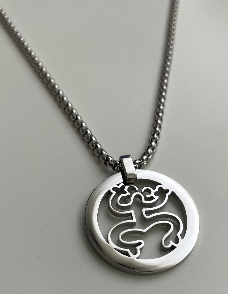 Puede incluir: Un collar de cadena de plata con un colgante redondo que presenta un dise&ntilde;o recortado de una rana estilizada.