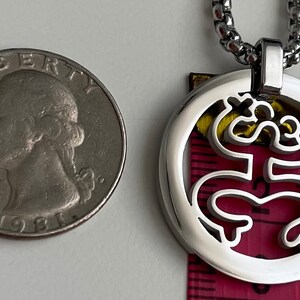 Puede incluir: Un collar de cadena de plata con un colgante redondo que presenta un dise&ntilde;o recortado de una rana estilizada. El colgante es aproximadamente del tama&ntilde;o de una moneda de 25 centavos de d&oacute;lar estadounidense.