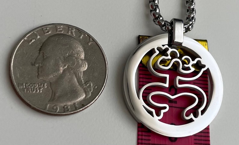 Puede incluir: Un collar de cadena de plata con un colgante redondo que presenta un dise&ntilde;o recortado de una rana estilizada. El colgante es aproximadamente del tama&ntilde;o de una moneda de 25 centavos de d&oacute;lar estadounidense.
