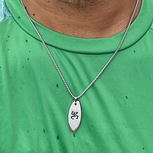 Op de afbeelding: Een zilveren ketting met een zilveren hanger in de vorm van een surfboard met het woord "ॐ" erop gegraveerd.