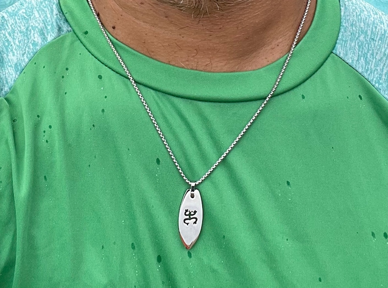 Op de afbeelding: Een zilveren ketting met een zilveren hanger in de vorm van een surfboard met het woord "ॐ" erop gegraveerd.