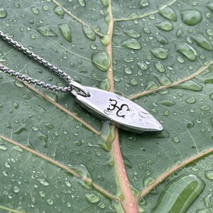 Op de afbeelding: Een zilveren ketting met een klein, ovaalvormig hanger. De hanger heeft een gegraveerd boomontwerp. De ketting ligt op een groen blad met waterdruppels.