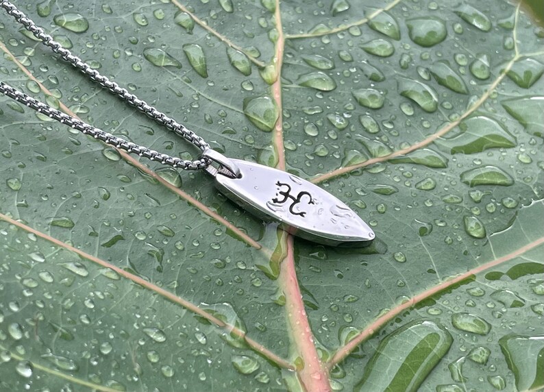 Op de afbeelding: Een zilveren ketting met een klein, ovaalvormig hanger. De hanger heeft een gegraveerd boomontwerp. De ketting ligt op een groen blad met waterdruppels.