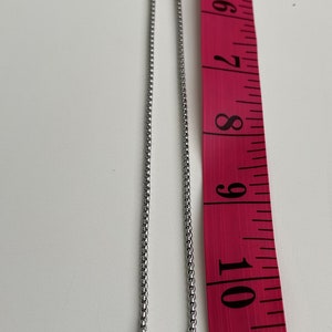 Op de afbeelding: Een zilveren ketting met een zilveren hanger met een uitgesneden ontwerp van een gestileerde figuur. De ketting wordt gemeten met een roze meetlint.