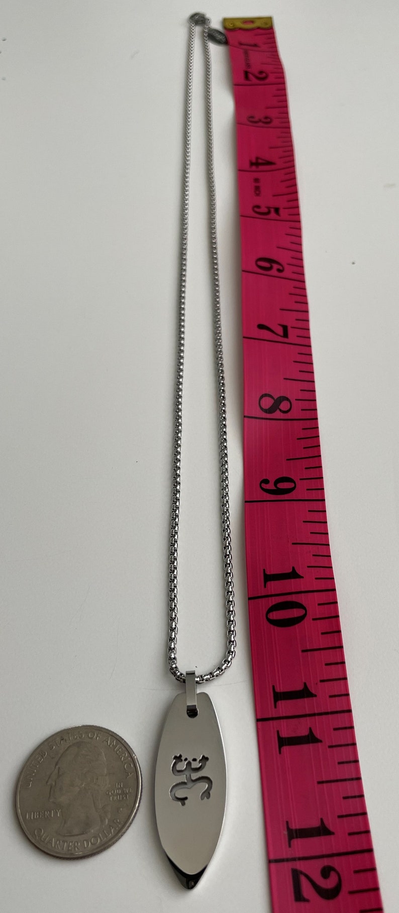 Op de afbeelding: Een zilveren ketting met een zilveren hanger met een uitgesneden ontwerp van een gestileerde figuur. De ketting wordt gemeten met een roze meetlint.