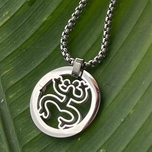 Puede incluir: Un collar de cadena de plata con un colgante redondo que presenta un dise&ntilde;o recortado de una rana estilizada. El colgante est&aacute; sobre un fondo de hojas verdes.