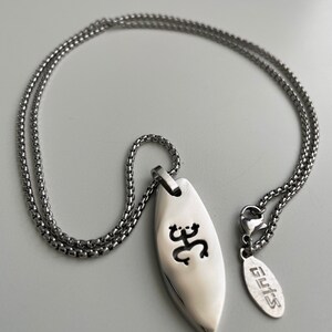 Op de afbeelding: Een zilveren ketting met een zilveren hanger met een gestileerd ontwerp. De hanger is surfboardvormig en heeft een uitgesneden ontwerp. Het label aan de ketting luidt "GUYS".