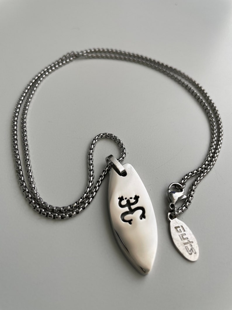 Op de afbeelding: Een zilveren ketting met een zilveren hanger met een gestileerd ontwerp. De hanger is surfboardvormig en heeft een uitgesneden ontwerp. Het label aan de ketting luidt "GUYS".