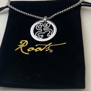 Puede incluir: Un collar de cadena de plata con un colgante de plata que presenta un dise&ntilde;o de rana estilizado. El colgante est&aacute; en una bolsa de terciopelo negro con la palabra "Roots" escrita en letras doradas.