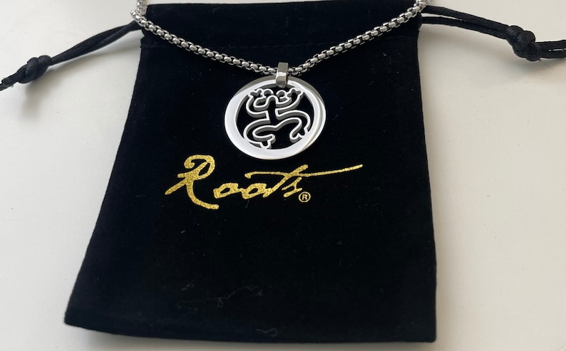 Puede incluir: Un collar de cadena de plata con un colgante de plata que presenta un dise&ntilde;o de rana estilizado. El colgante est&aacute; en una bolsa de terciopelo negro con la palabra "Roots" escrita en letras doradas.