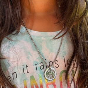 Puede incluir: Un collar de cadena de plata con un colgante redondo que presenta un dise&ntilde;o recortado de una rana. El collar lo lleva una persona que lleva una camisa azul claro con un patr&oacute;n de arco&iacute;ris y el texto "Cuando llueve busca arco&iacute;ris".