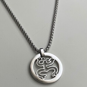 Puede incluir: Un collar de cadena de plata con un colgante redondo que presenta un dise&ntilde;o recortado de una rana estilizada.