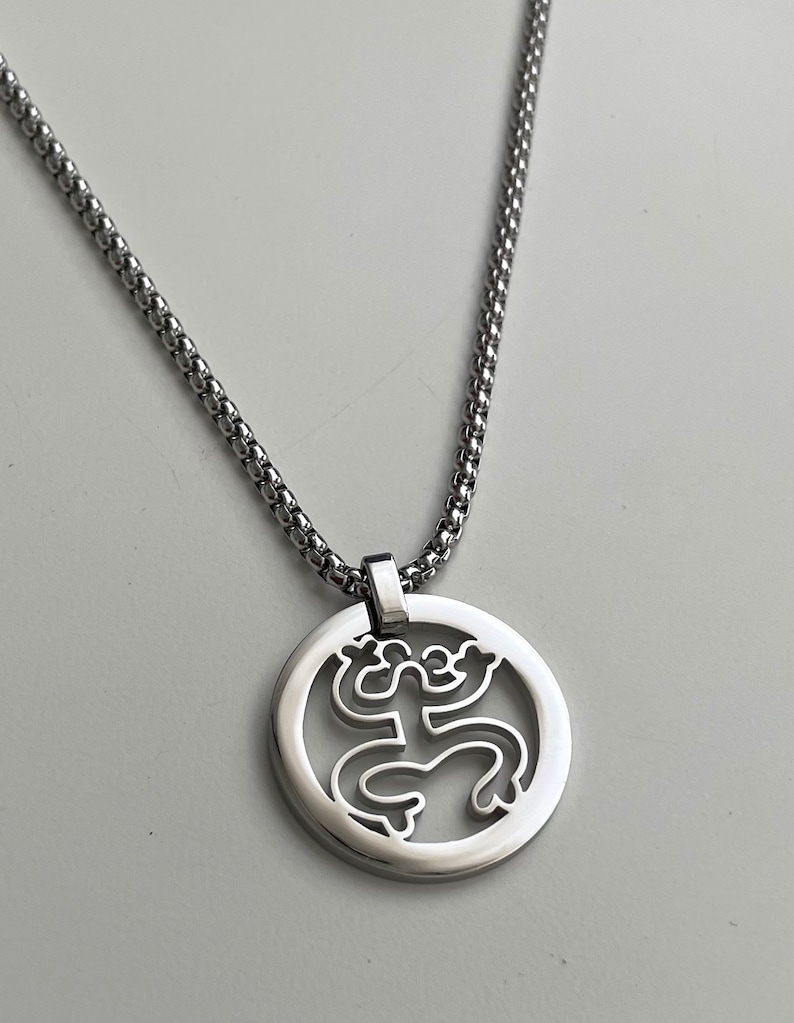 Puede incluir: Un collar de cadena de plata con un colgante redondo que presenta un dise&ntilde;o recortado de una rana estilizada.