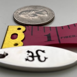 Op de afbeelding: Een zilveren ovale hanger met een zwarte inscriptie van de letters "3C" aan een zilveren ketting. De hanger ligt naast een roze meetlint met het getal "1" zichtbaar en de woorden "FIBER-GLASS" erop gedrukt.