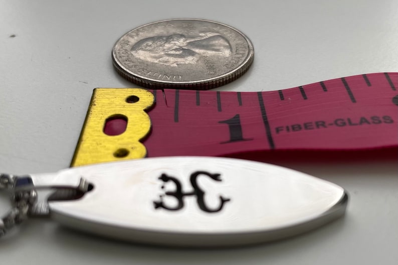 Op de afbeelding: Een zilveren ovale hanger met een zwarte inscriptie van de letters "3C" aan een zilveren ketting. De hanger ligt naast een roze meetlint met het getal "1" zichtbaar en de woorden "FIBER-GLASS" erop gedrukt.