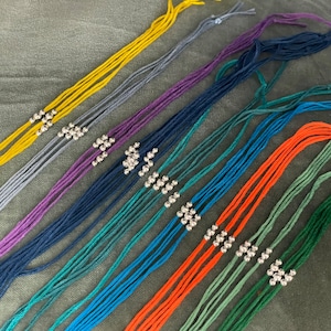 Puede incluir: Un conjunto de nueve pulseras de cuerda de colores vivos con cuentas plateadas. Las pulseras están dispuestas en fila, con la pulsera amarilla en la parte superior y la pulsera verde en la parte inferior.