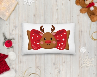 May include: A rectangular white pillow featuring a cartoon reindeer design. The reindeer has a brown face, antlers, and a red nose. A large red bow with white polka dots frames the reindeer's face. The pillow is a festive holiday decoration.
