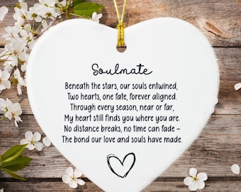 May include: White heart-shaped ornament with the word "Soulmate" and a poem printed in black. A gold string is attached for hanging. The background is a rustic wooden surface with white flowers.