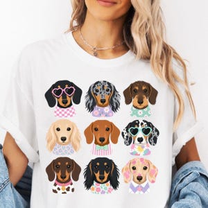 Puede incluir: Una camiseta blanca con un estampado gráfico de nueve perros salchicha dispuestos en cuadrícula. Cada perro tiene un pelaje y color únicos, y algunos llevan accesorios como gafas de sol en forma de corazón o pajaritas.