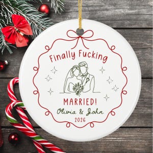 May include: A round white ceramic ornament featuring a red decorative border and a gold hanging string. The text reads Finally Fucking MARRIED! Olivia & John 2026, accompanied by a black line art illustration of a bride and groom.