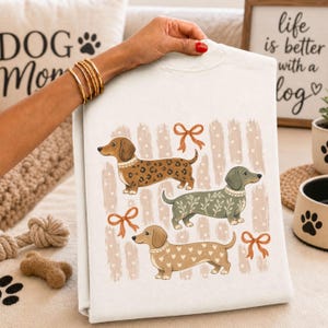 Può includere: Una felpa bianca con una stampa grafica di tre cani bassotti. Il cane in alto ha un motivo leopardato marrone, quello centrale è verde salvia con foglie bianche e quello in basso è color marrone chiaro con cuori bianchi. Tre fiocchi arancioni.
