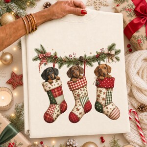 May include: A cream-colored sweatshirt featuring a graphic of three brown dachshunds peeking out of patterned Christmas stockings. The stockings hang from a green pine garland decorated with red berries and bows, creating a festive holiday scene.