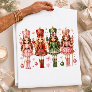 Op de afbeelding: Een wit sweatshirt met een grafische print van vier cartoon notenkrakerfiguren. De figuren dragen feestelijke uniformen in roze, rode en groene tinten met gouden details en zijn omringd door kleine sterretjes, strikjes en kerstballen.