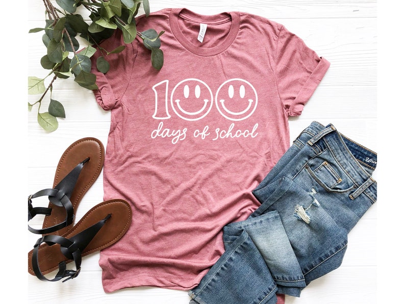 May include: A maroon t-shirt with the text "100 days of school" in white, featuring two smiley faces. The shirt is paired with blue jeans and black sandals with brown soles. Green leafy sprigs are also in the image.