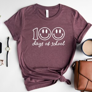 May include: Burgundy t-shirt with the text "100 days of school" and two smiley faces in white. The shirt is displayed on a white surface with accessories, including a watch, glasses, and a purse.